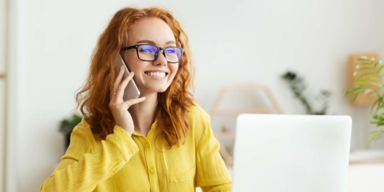 Smart-working Immagine di una ragazza con occhiali sorridente mentre risponde al cellulare davanti a un computer portatile aperto di fronte a lei